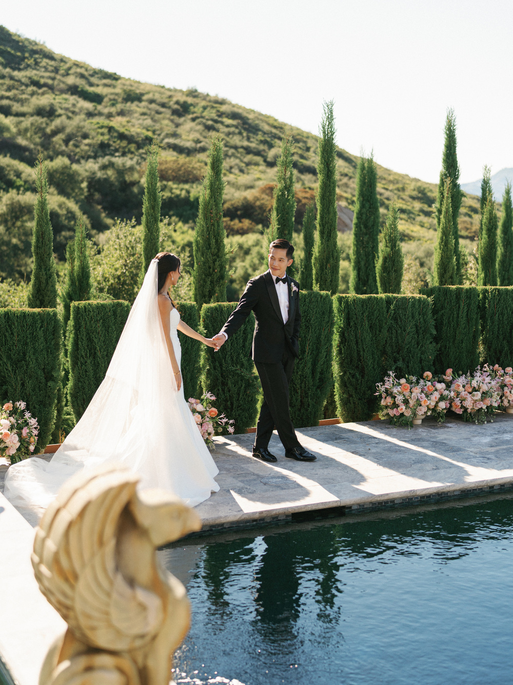 Bride and groom holding hands by a pool with cypress trees and hills