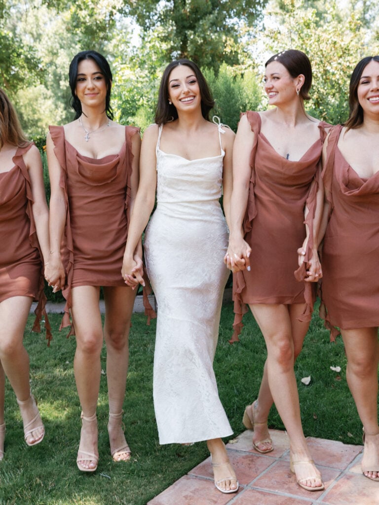 Contemporary bridal party style featuring bridesmaids in terracotta cowl neck mini dresses walking hand-in-hand.