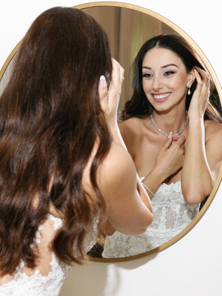 Glamorous bride adjusting fine diamond jewelry in a round mirror during her bridal getting ready session.