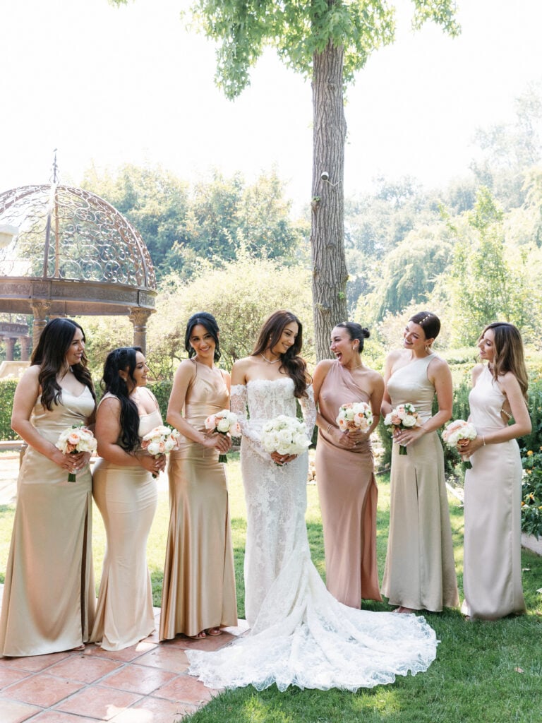 Bride in lace gown posing with bridesmaids in champagne silk dresses during morning wedding coverage hours.