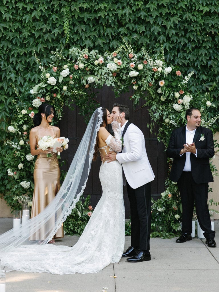 Romantic first kiss at an outdoor garden altar framed by a lush, circular white and blush floral arch.