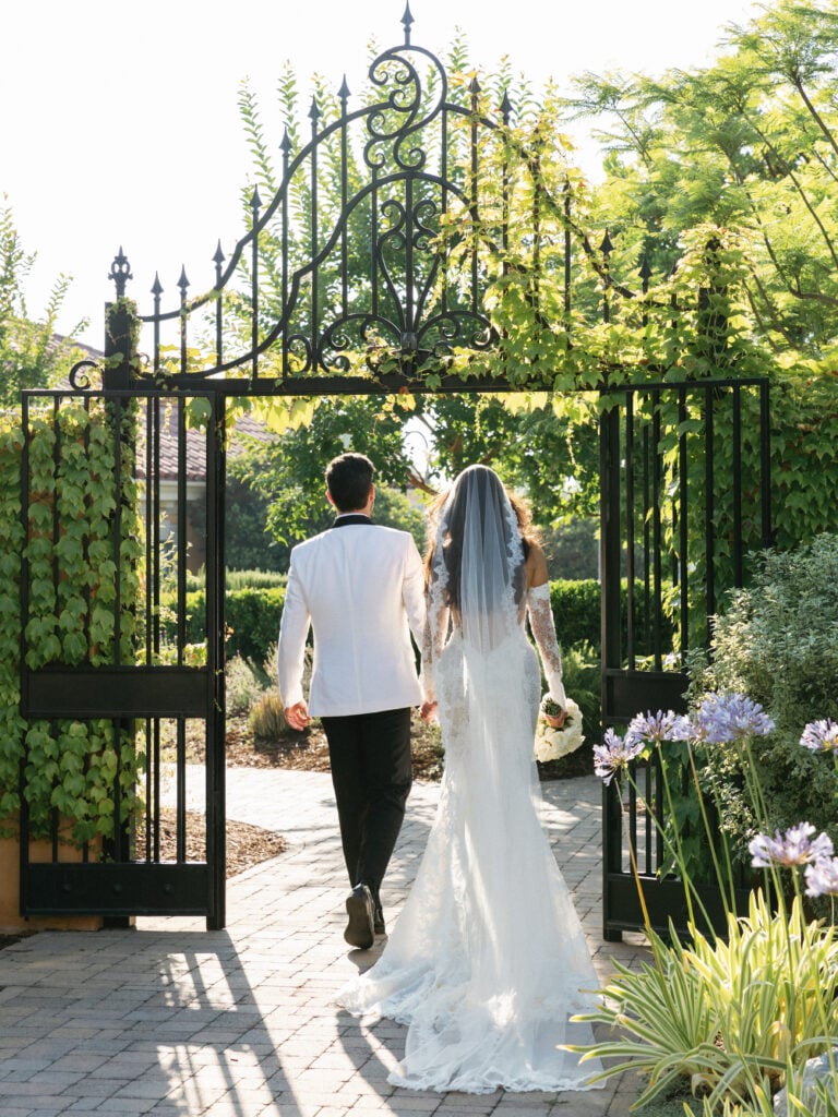 Timeless rear-view portrait of newlyweds walking through ornate wrought iron gates at a European-inspired estate.