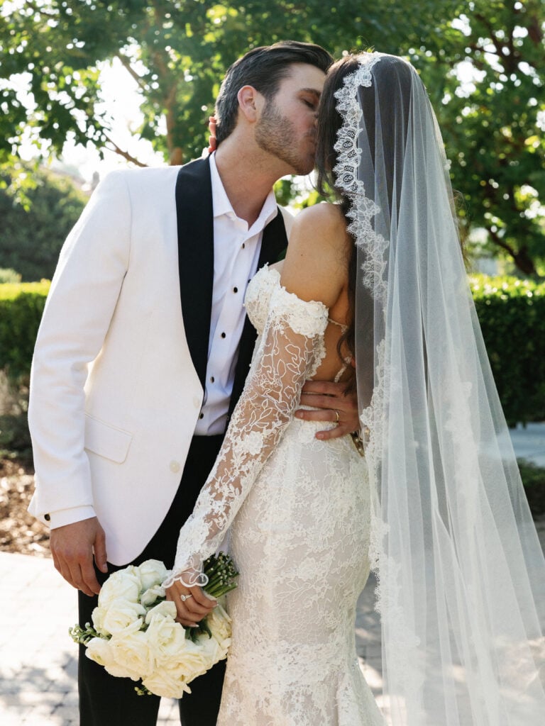 Intimate newlywed kiss showcasing a sheer lace off-the-shoulder sleeve trend and a timeless cathedral veil.