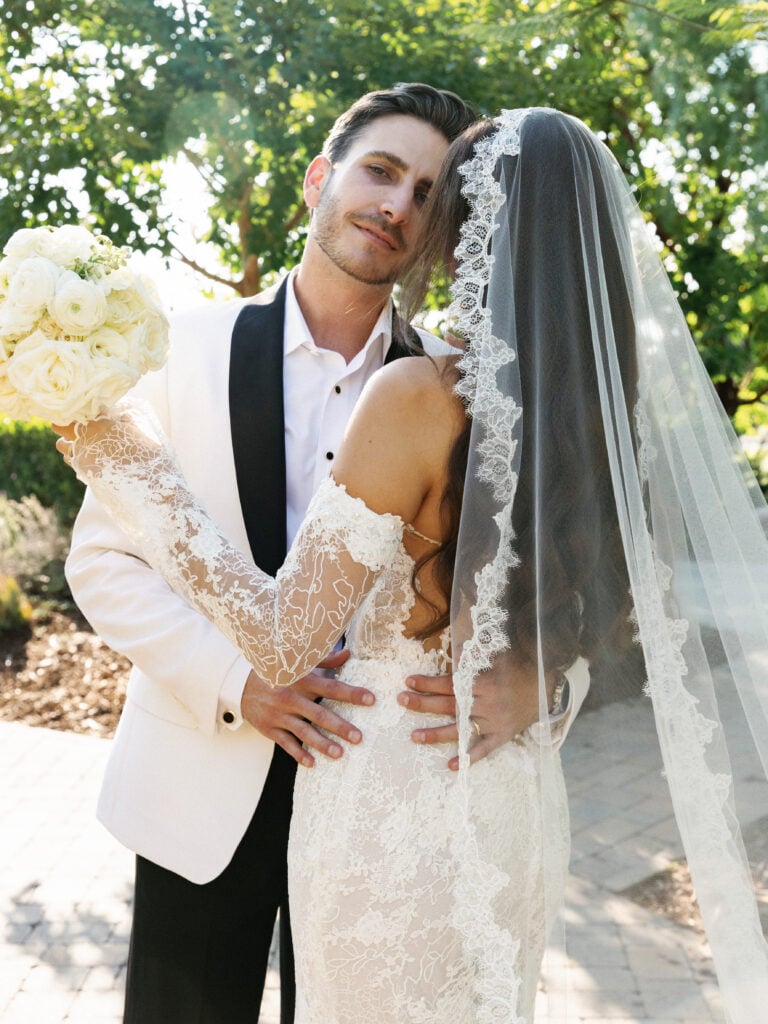 Editorial close-up wedding portrait showcasing a dramatic lace mantilla veil trend and modern white tuxedo style.