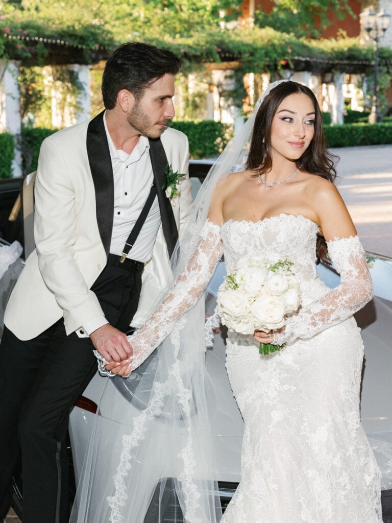 Editorial style bride and groom posing by a luxury getaway car for their wedding day portraits.
