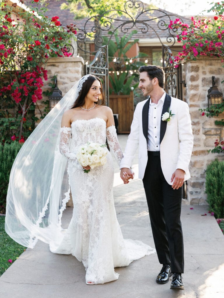 Bride in lace couture gown and groom in white tuxedo jacket walking hand in hand for outdoor wedding portraits.