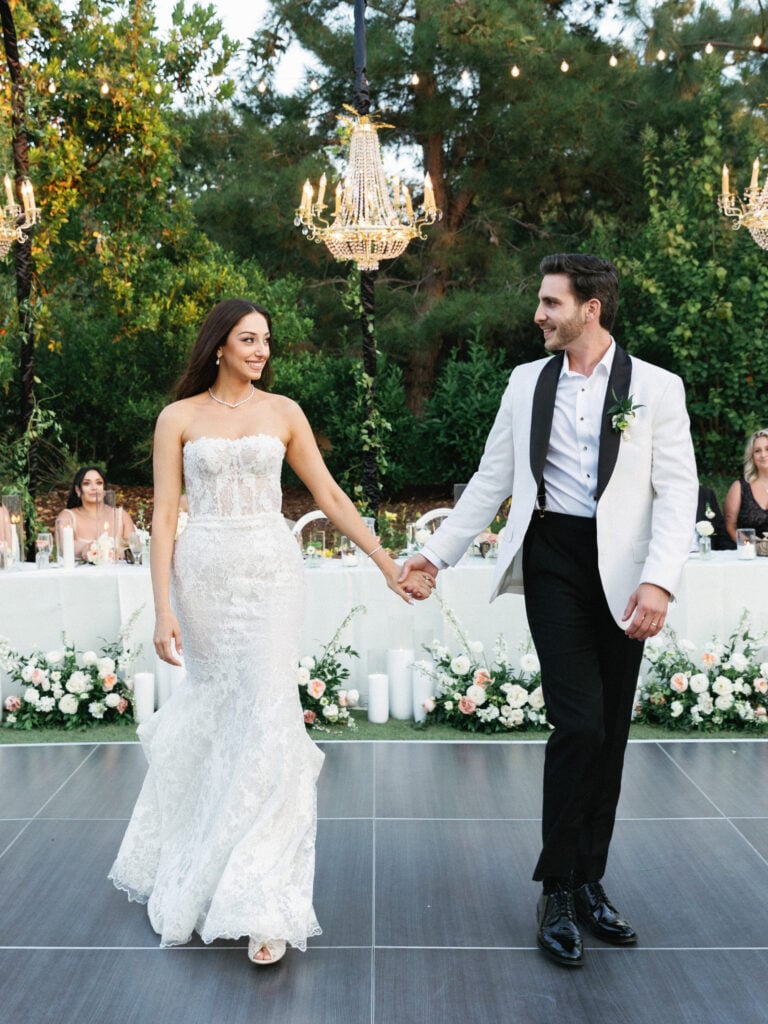 Newlyweds walking onto a modern outdoor dance floor under hanging crystal chandeliers at a black-tie reception.