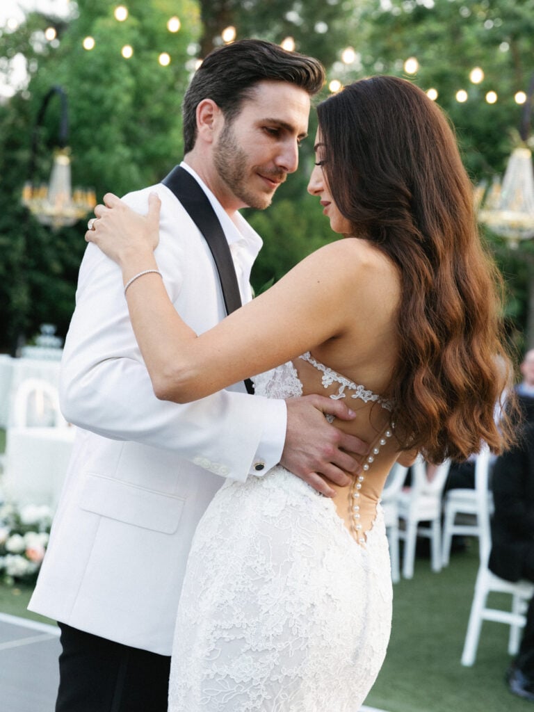Elegant couple sharing an intimate first dance at an outdoor twilight wedding reception with string lights.