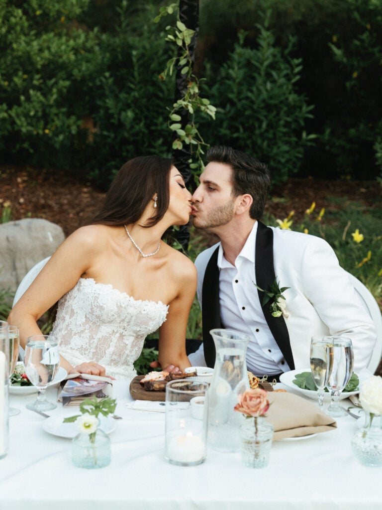 Candid bride and groom kiss at an alfresco sweetheart table during golden hour wedding coverage hours.