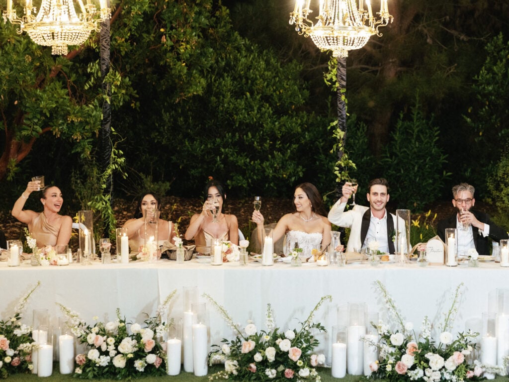Glamorous alfresco wedding head table design featuring cascading ground florals, pillar candles, and hanging crystal chandeliers.