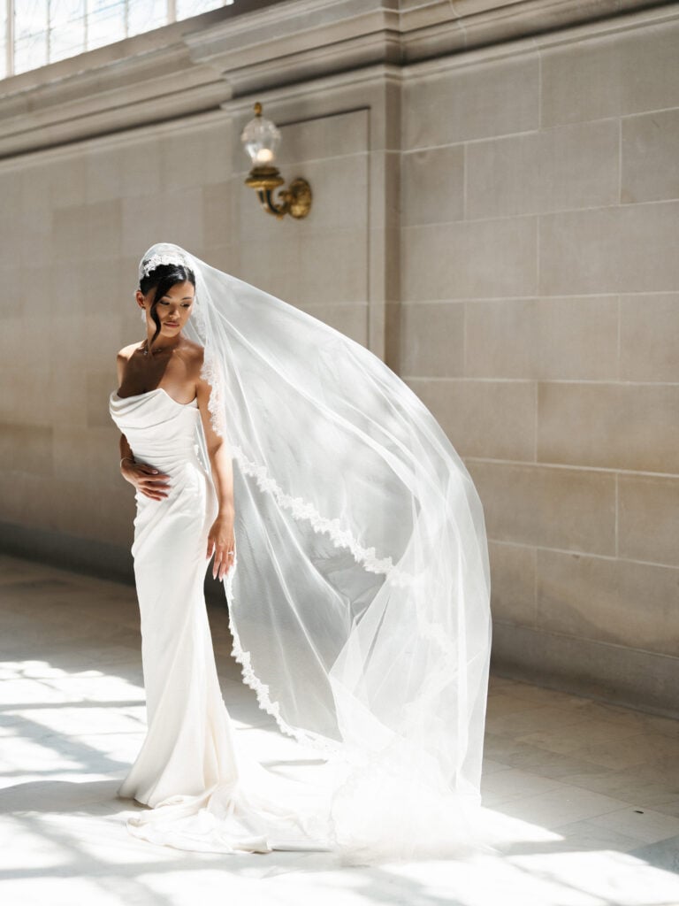 Dramatic indoor wedding dress shot using harsh window light to illuminate a flying veil for high-end wedding venue photography.