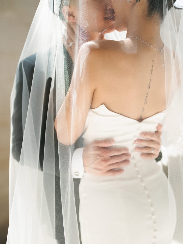 Romantic under-the-veil embrace showcasing modern strapless wedding dress back details with delicate buttons and a classic mantilla veil.