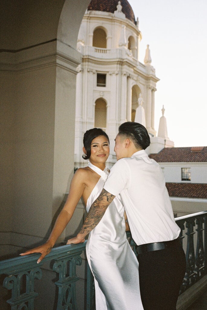 Candid film wedding photography of an elegant couple embracing on a historic balcony with a scenic city hall dome backdrop.