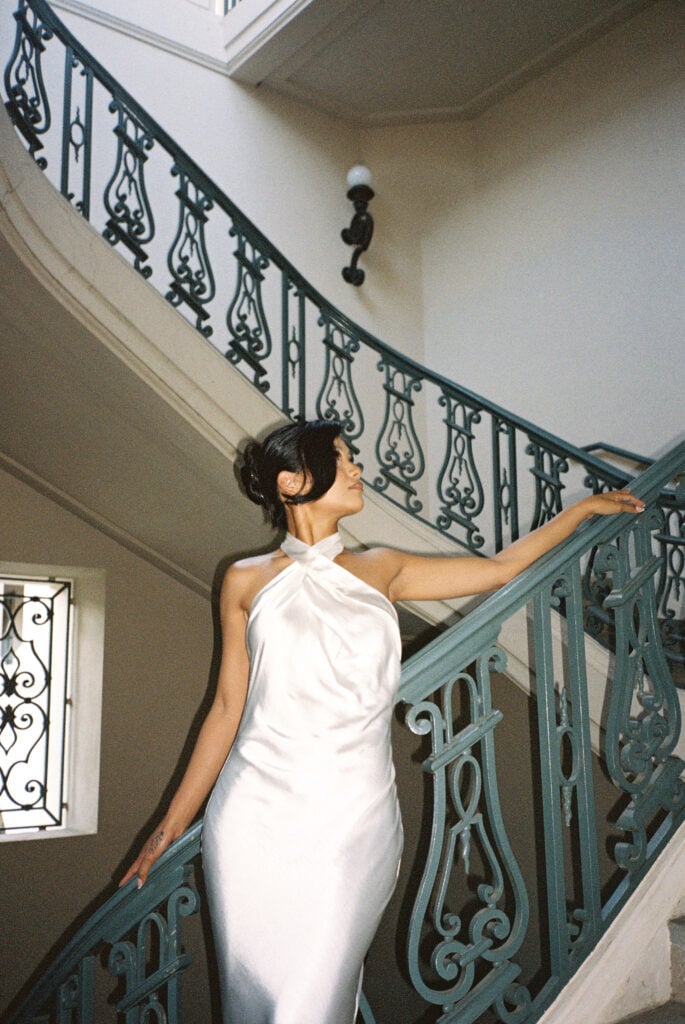 Editorial film wedding portrait of a chic bride in a minimalist silk halter dress posing on a vintage architectural staircase.