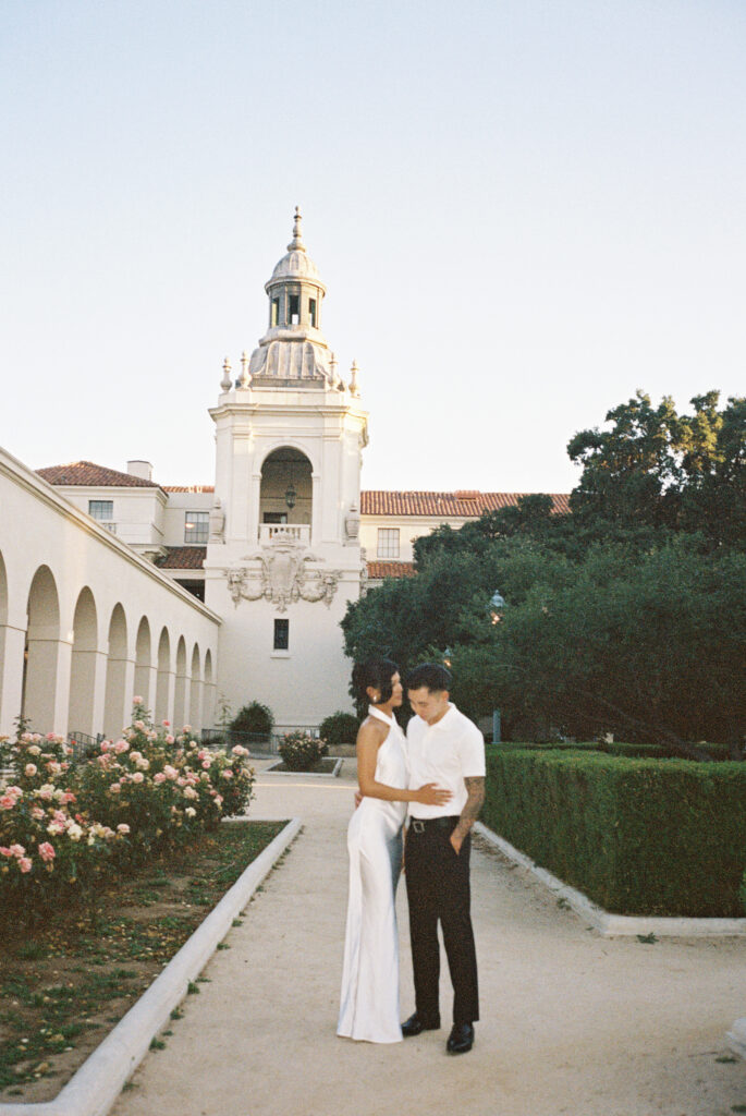 Editorial film wedding portrait of a stylish couple standing in a romantic garden courtyard with a grand Spanish-style bell tower backdrop.