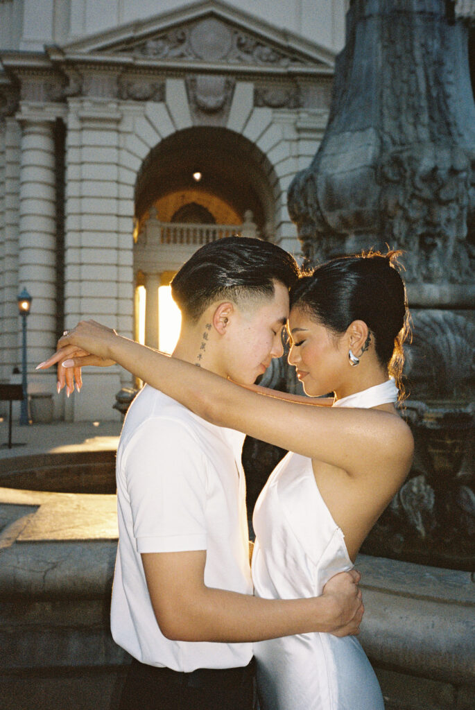 Romantic golden hour film wedding photography of an elegant couple embracing tenderly near a historic stone fountain.