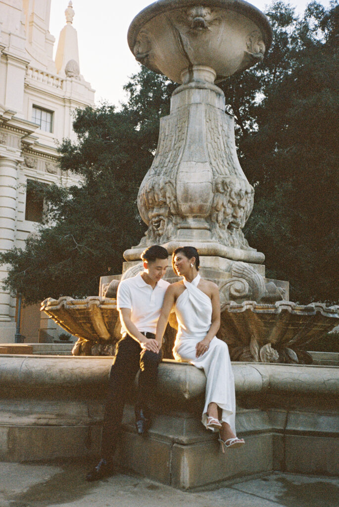 Editorial film wedding portrait of a chic couple sitting on a vintage sculptural fountain, capturing an effortless European-style elopement.