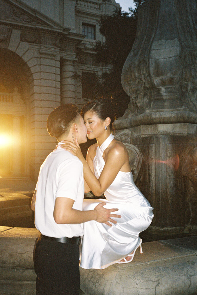 Editorial film wedding portrait of a chic bride in a glowing silk halter gown kissing her groom near a historic fountain.