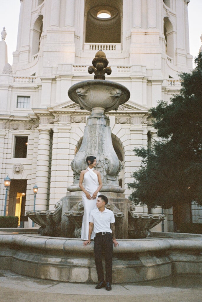 Grand film wedding portrait of an elegant couple posing by a historic stone fountain with a majestic city hall backdrop.