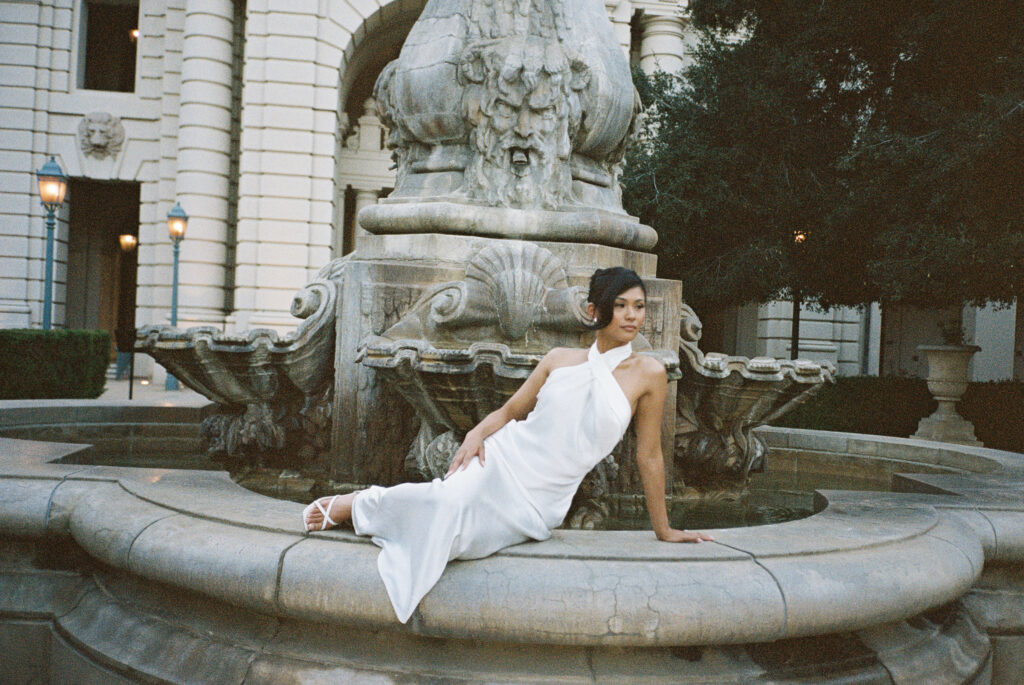 Editorial film wedding photography featuring a modern bride in a silk halter gown and strappy sandals reclining by a vintage fountain.