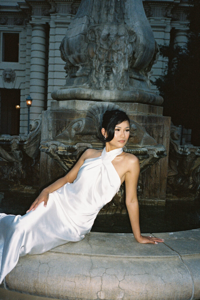 High-fashion flash film bridal portrait of a chic bride in a white silk halter gown posing by a stone fountain at dusk.