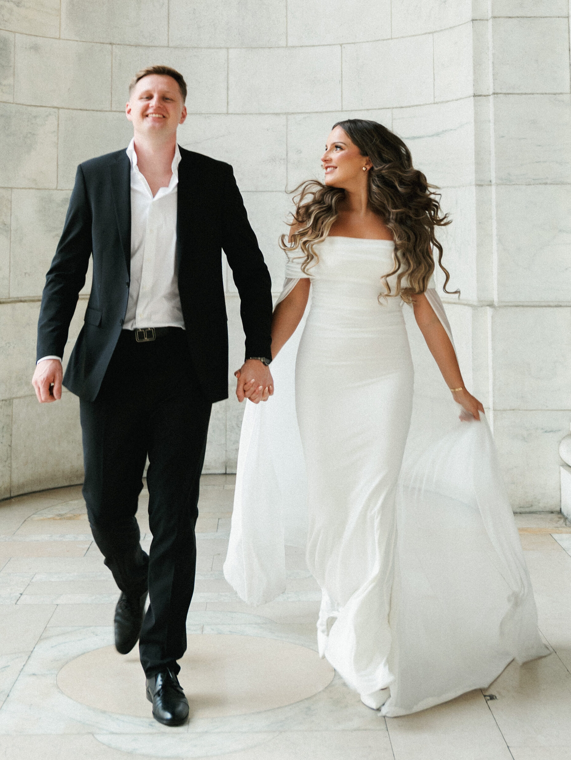 Bride in off-the-shoulder gown with cape sleeves and groom walking hand-in-hand inside a marble rotunda.