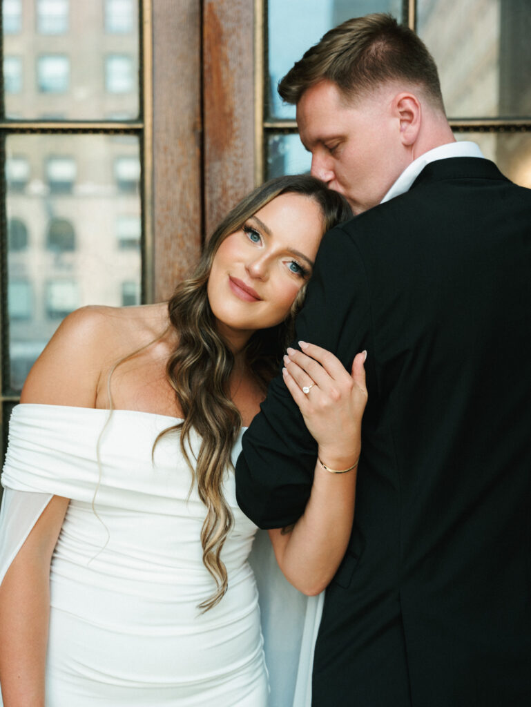 Close-up of bride leaning against groom while he kisses her forehead beside a glass window.