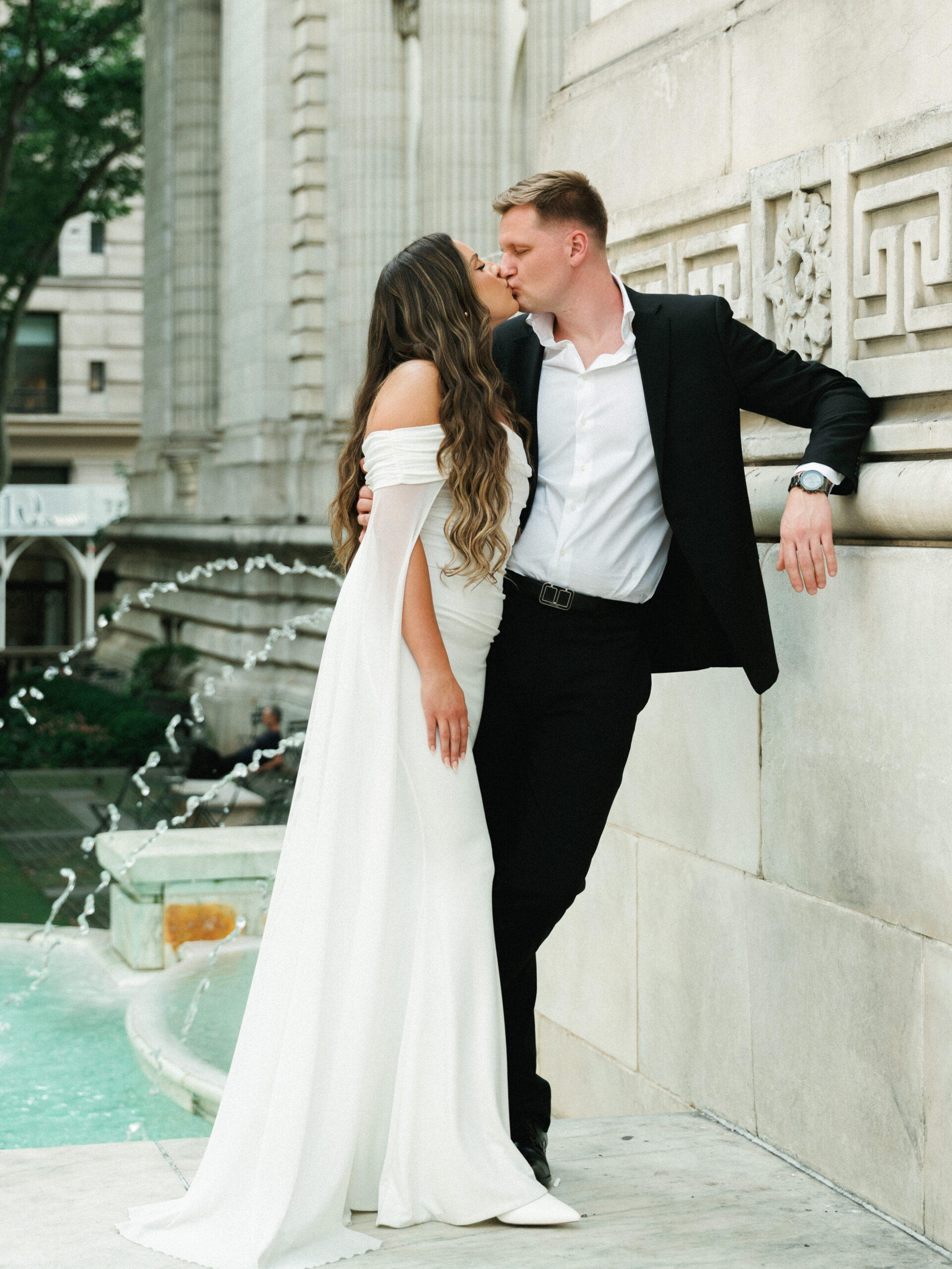 Bride and groom kissing beside an outdoor stone fountain against a carved classical building wall.