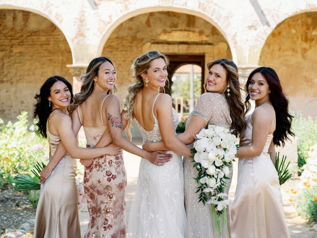 Smiling bride with her bridesmaids in champagne satin and sequin dresses looking over their shoulders, capturing a candid group moment for editorial wedding photography.