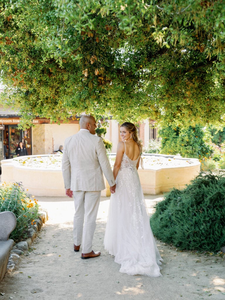 Candid bride in a sparkly tulle gown walking hand-in-hand with her groom past a stone fountain under lush green trees, highlighting relaxed editorial wedding photography.