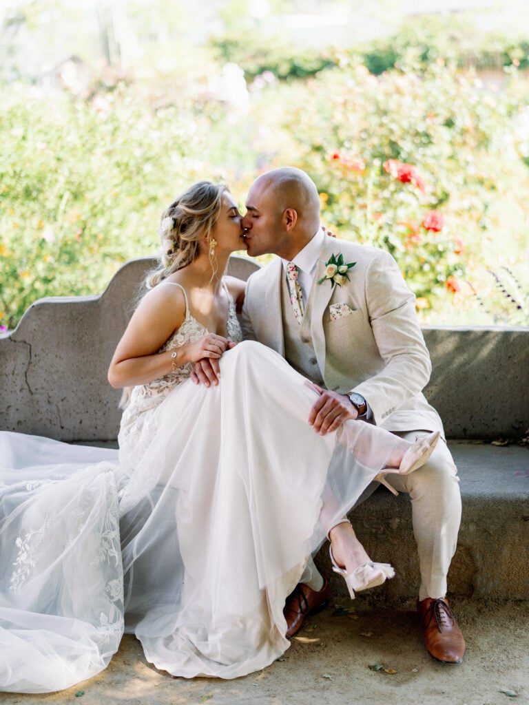 Romantic bride and groom sharing a passionate kiss on an outdoor stone bench in a blooming garden, capturing natural movement for modern editorial wedding photography.