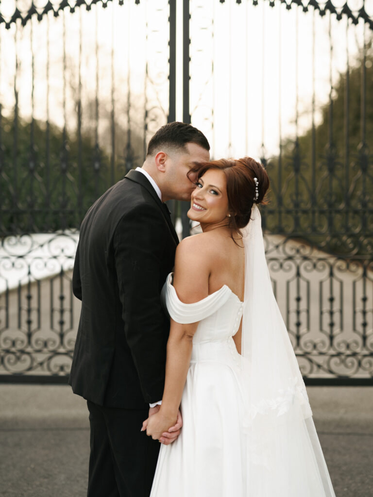 Smiling bride in an off-the-shoulder satin ballgown looking over her shoulder as the groom kisses her cheek before tall ornate wrought-iron gates, showcasing relaxed editorial wedding photography.