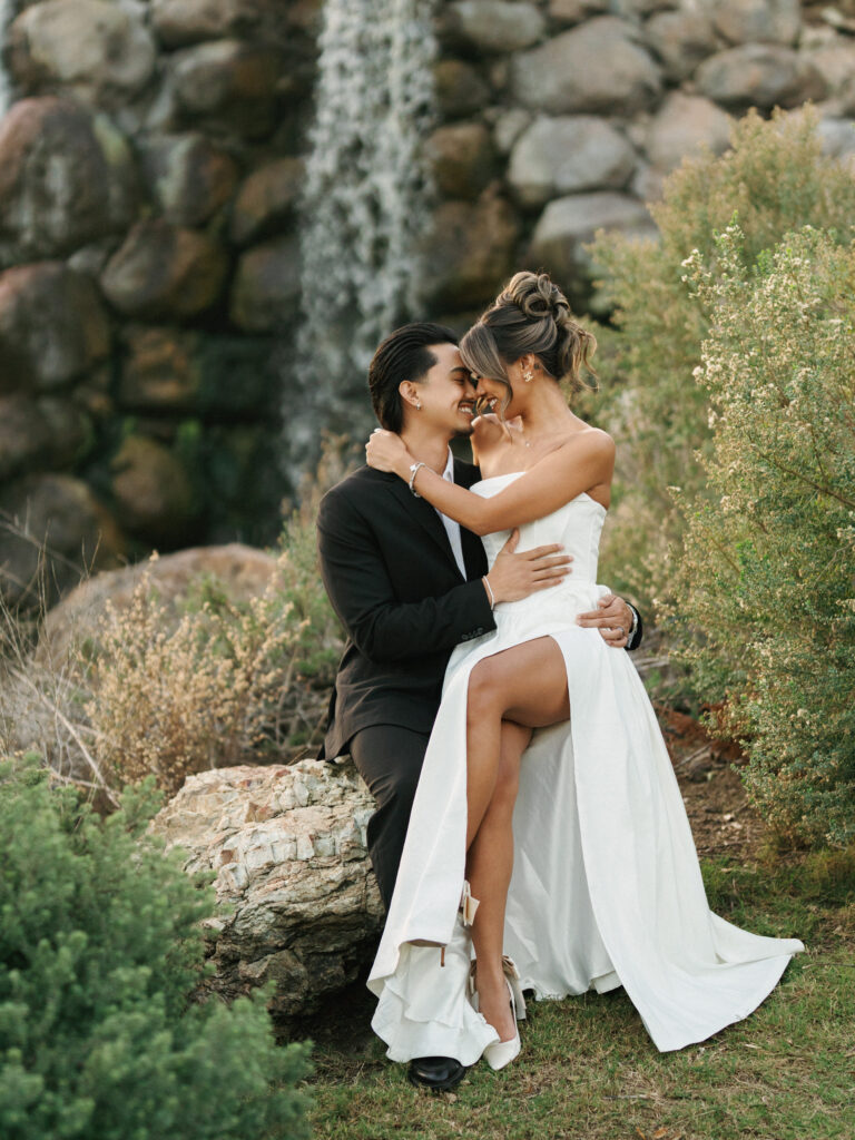 Candid bride in a strapless high-slit wedding gown laughing with her groom on a rock near a waterfall, illustrating a relaxed approach to editorial wedding photography.