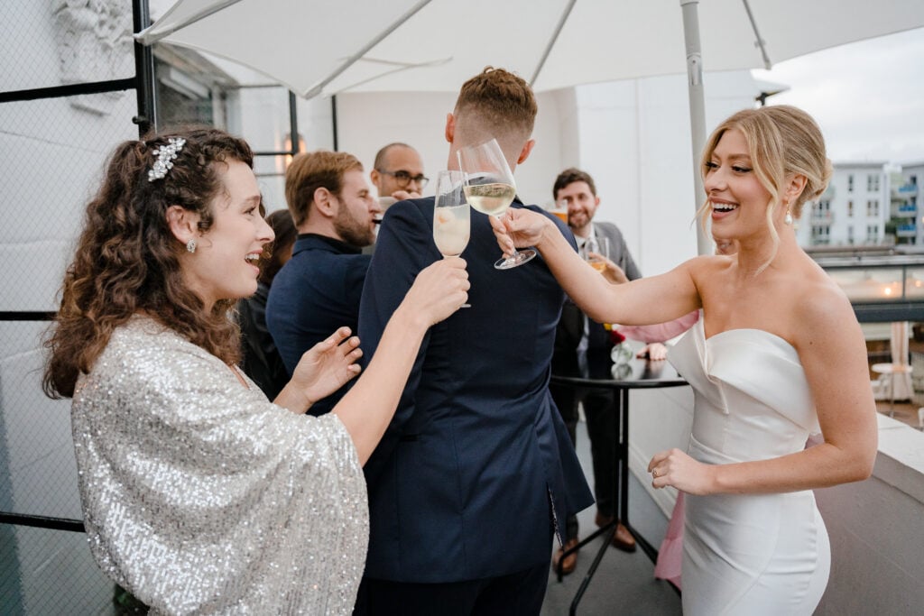 Smiling bride in a strapless architectural sweetheart gown clinking wine glasses with guests at a chic rooftop welcome party during her wedding weekend coverage.