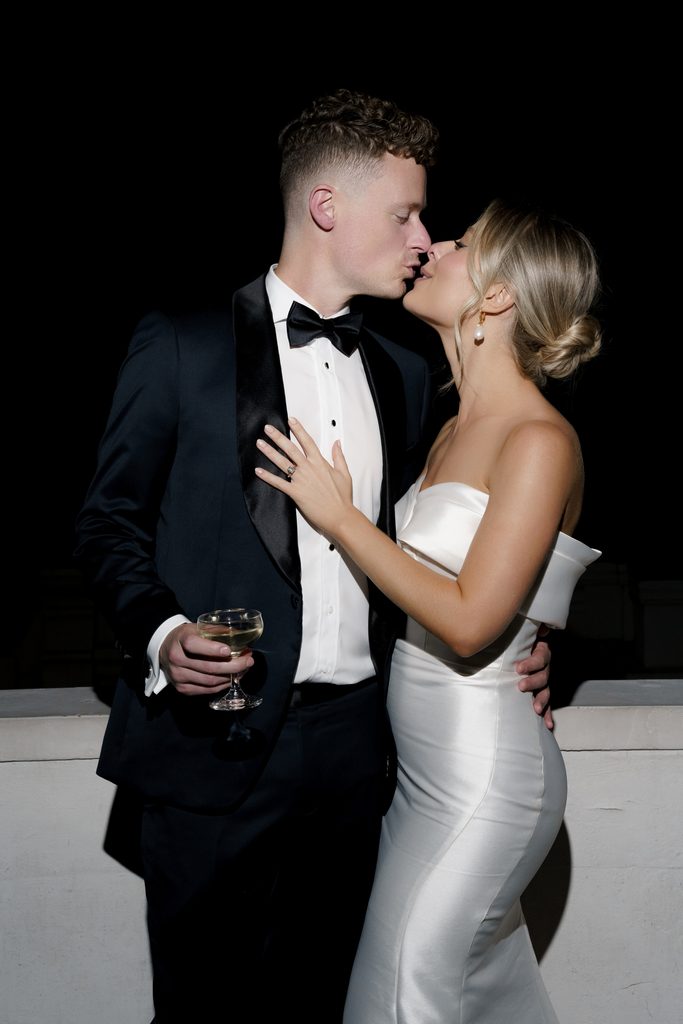Chic reception wedding photo ideas capturing a black-tie couple kissing while holding a champagne coupe at night.