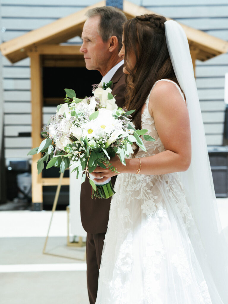 Bride with a long veil and white bouquet walking down the aisle with her father in a brown suit