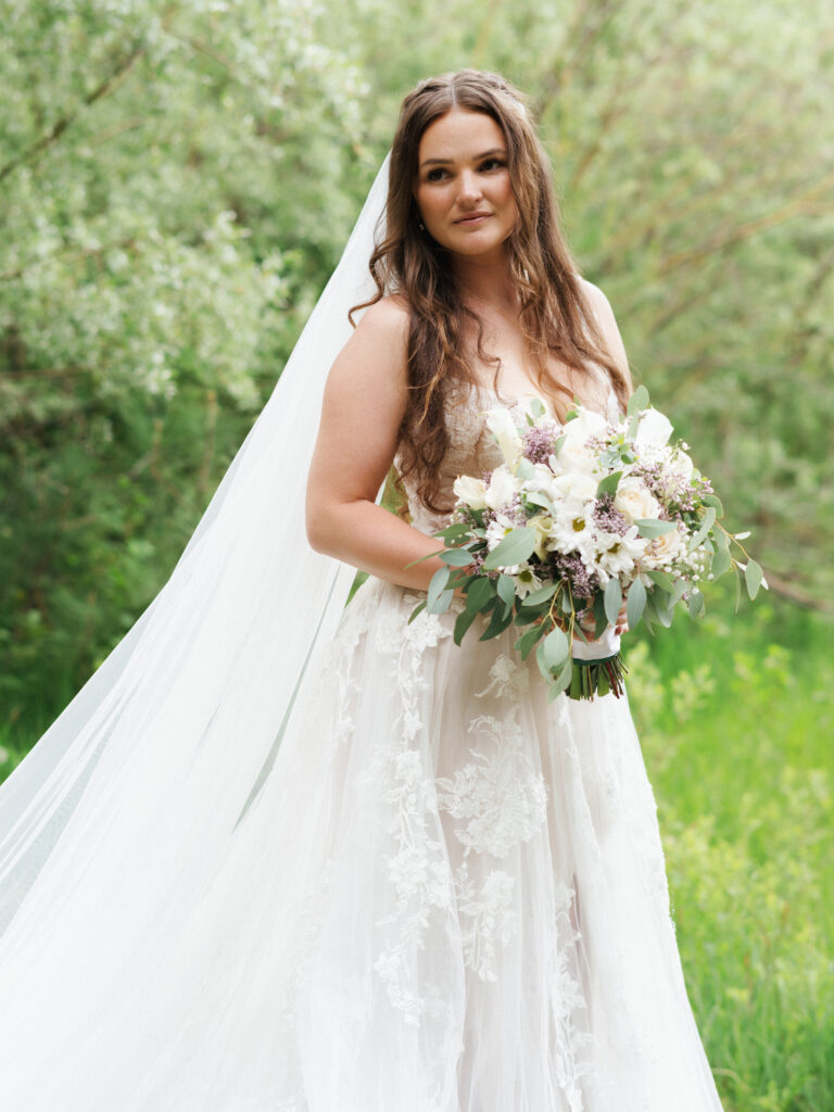 Bride with a long veil holding a white and purple bouquet on a green meadow background