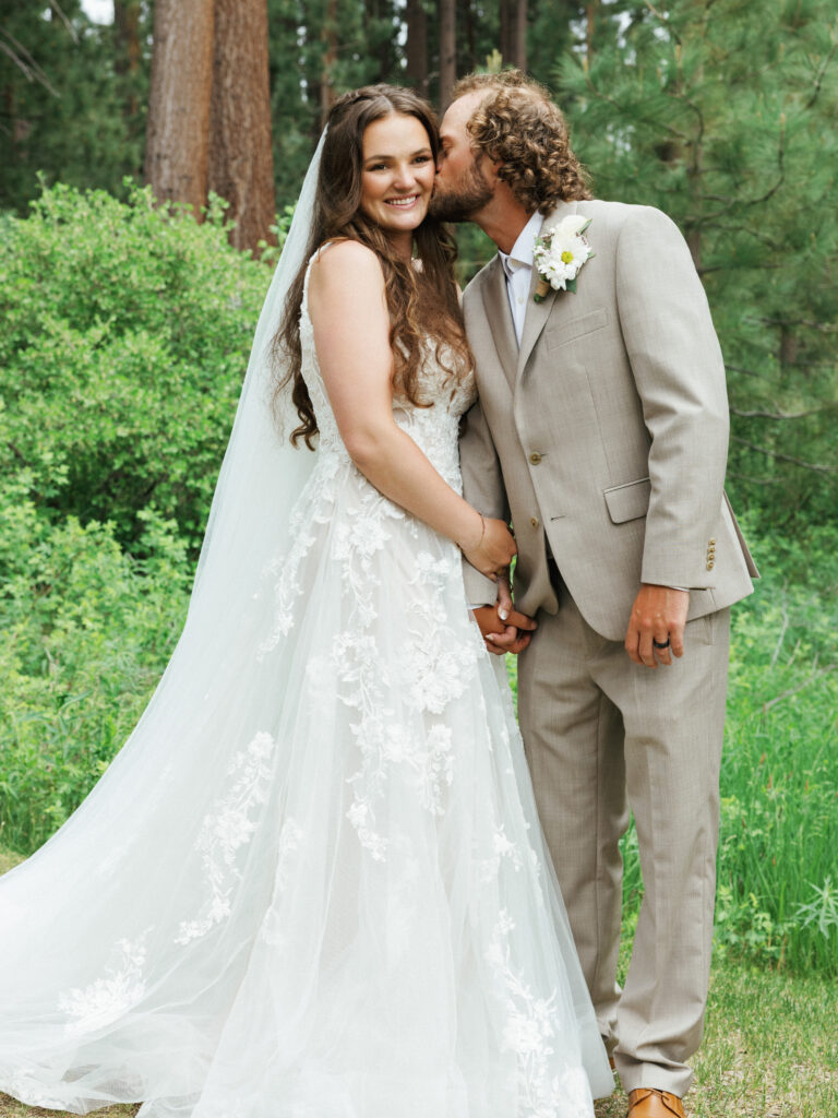 Groom kissing his smiling bride on the cheek while standing in a pine forest grove
