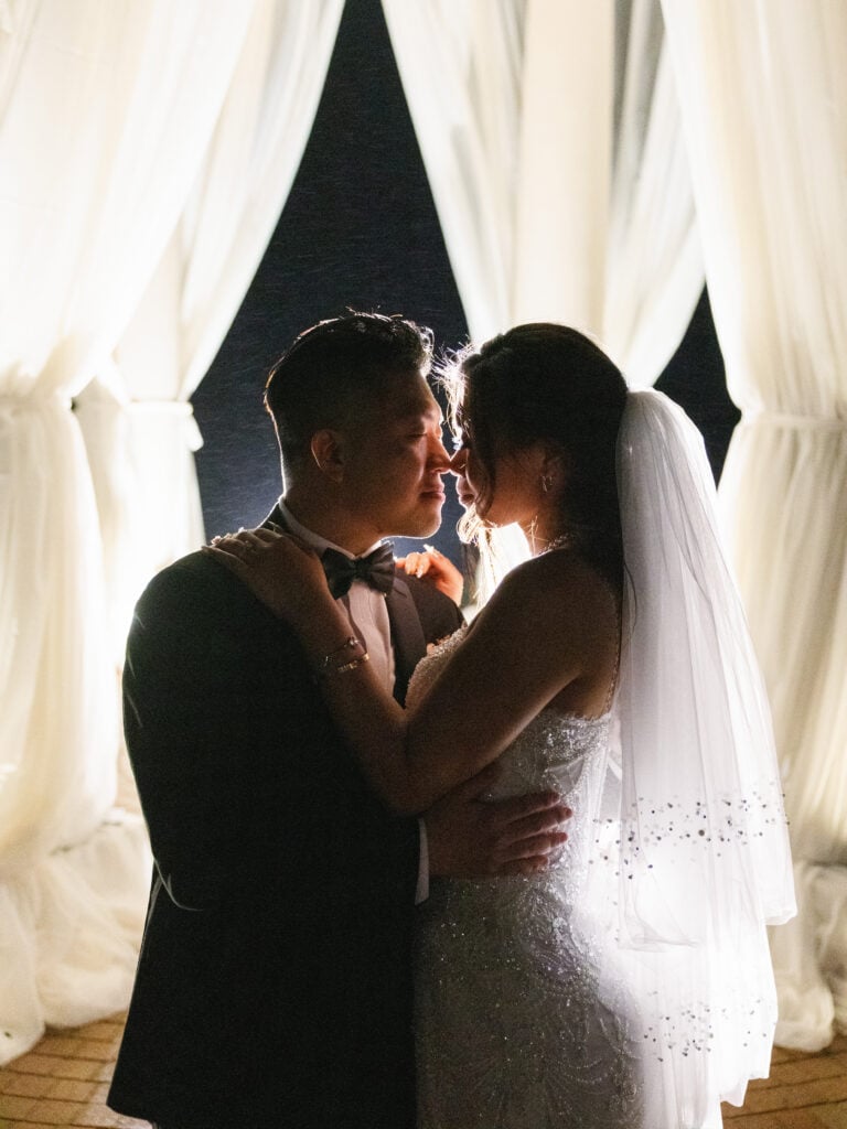Intimate back-lit newlywed portrait during a romantic night drizzle, showcasing dramatic couture details under a misty veil.