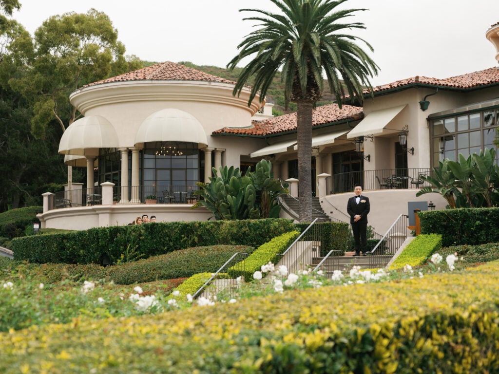 Classic black-tie groom standing by the elegant Mediterranean-style villa at The Resort at Pelican Hill under a soft, overcast sky.