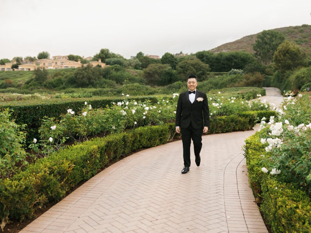 Dapper groom walking along a lush garden path, demonstrating a proactive wedding rain plan for outdoor portrait sessions.