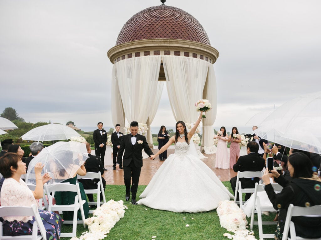 Joyful newlyweds outdoor recessional at The Resort at Pelican Hill rotunda, demonstrating a stylish wedding rain plan with clear umbrellas.