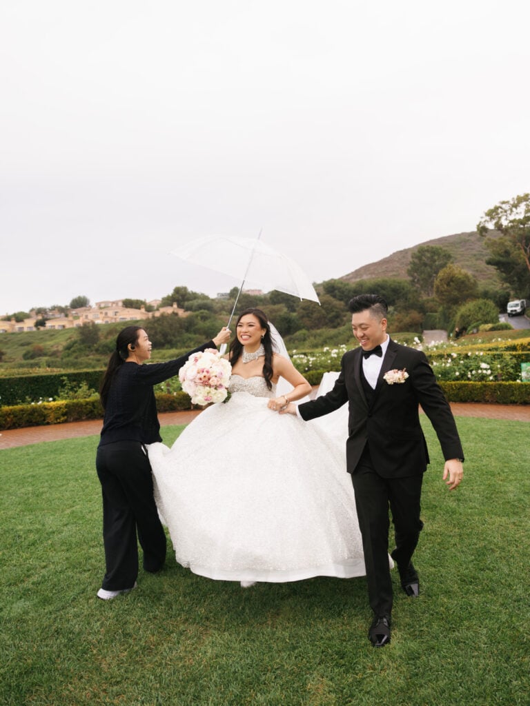 Helpful wedding coordinator assisting the bride and groom with a white umbrella, showing a seamless wedding rain plan execution in Newport Beach.