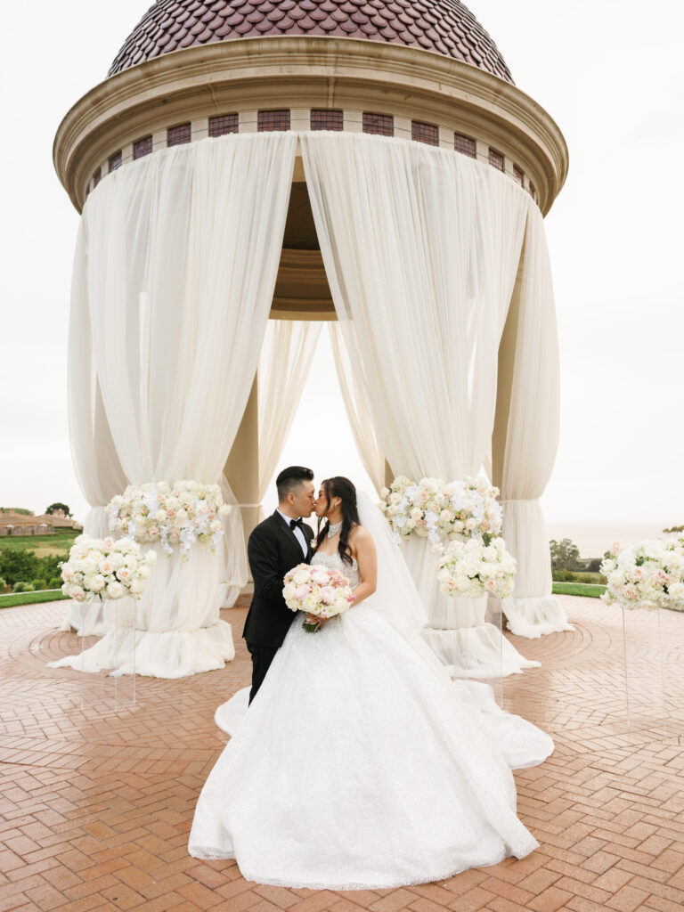 Romantic newlyweds kiss framed by lush white orchid altar arrangements, utilizing a smart wedding rain plan for flawless overcast lighting.