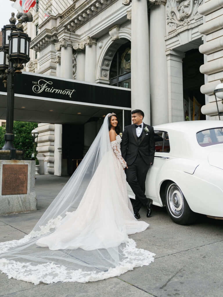 Elegant bride in a lace-sleeve wedding dress with a dramatic cathedral veil and a groom in a classic tuxedo posing by a vintage white getaway car outside The Fairmont San Francisco hotel, capturing timeless editorial wedding photography.