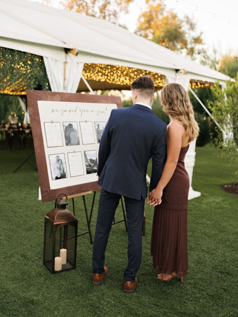 Chic wedding guests looking at an elegant seating chart with black and white photos next to a luxury reception tent during wedding weekend coverage.