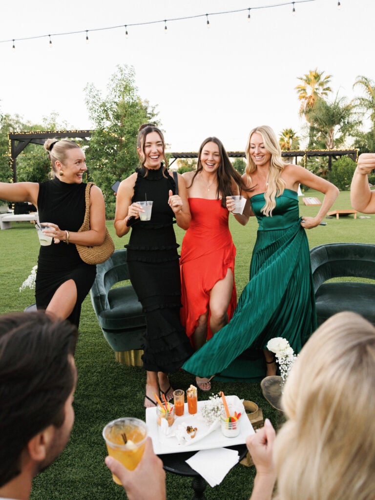 Lively female guests in colorful cocktail dresses dancing and enjoying signature cocktails on a green lawn, highlighting vibrant wedding weekend coverage.