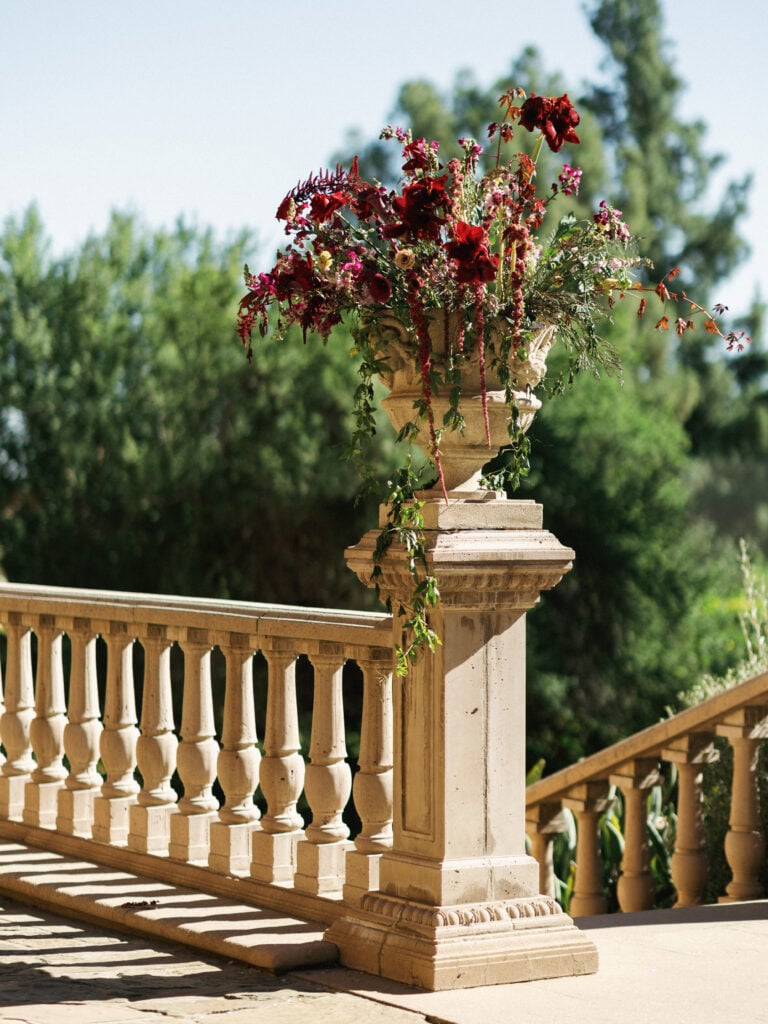 Luxury burgundy and rose floral arrangement in a stone urn on a classical balustrade at a California estate, perfect inspiration for high-end destination wedding travel.