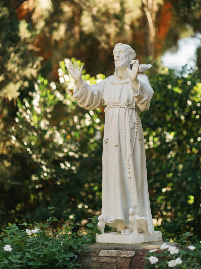 Elegant St. Francis stone statue surrounded by lush greenery and white flowers in the gardens of a historic California villa during a luxury destination wedding travel production.