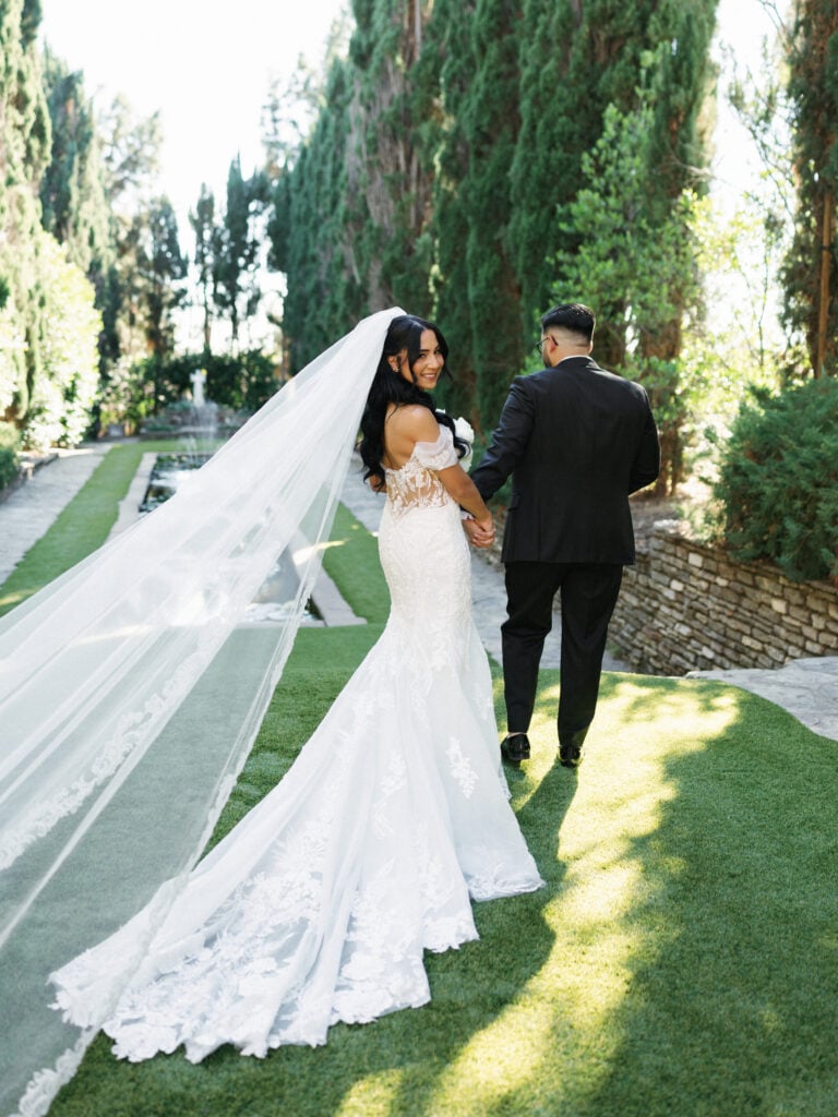 Elegant bride in an off-the-shoulder lace wedding dress with a long train and cathedral veil walking with her groom during their destination wedding travel.
